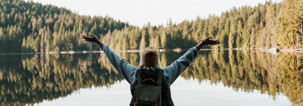 Eine junge Digitalnomadin steht vor einem See und freut sich.