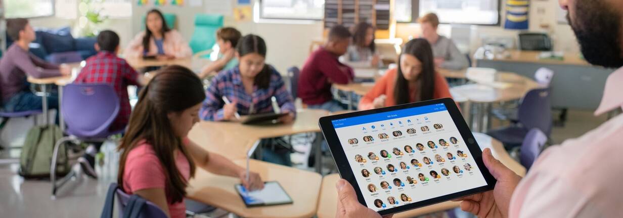 Lehrperson steht vor der Klasse mit iPad, das alle Lernenden zeigt